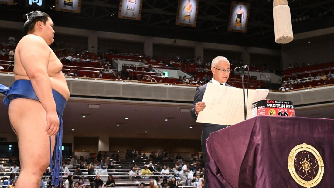 大相撲の会場にて、K&K PROTEIN BEEFが置かれた演台で表彰状を読み上げる男性と、それを見守る力士の様子の写真