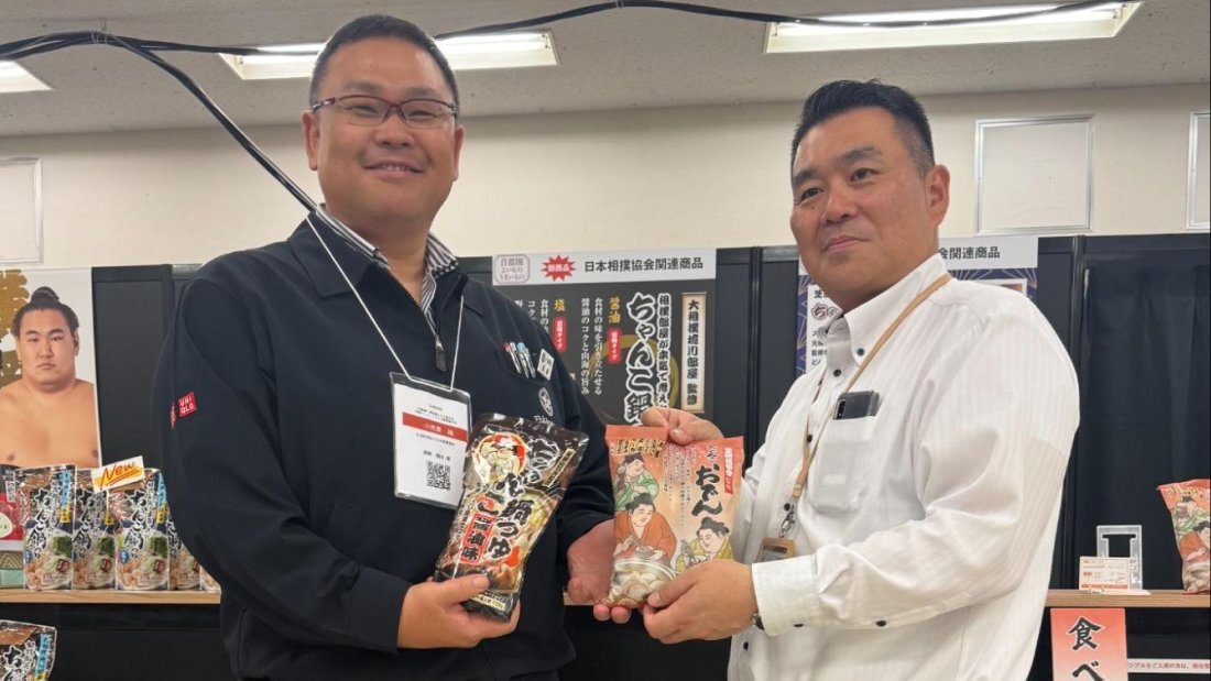 日本相撲協会関連商品である「ちゃんこ鍋つゆ」と「おでん」を手に持ち笑顔で並ぶ二人のスタッフの写真